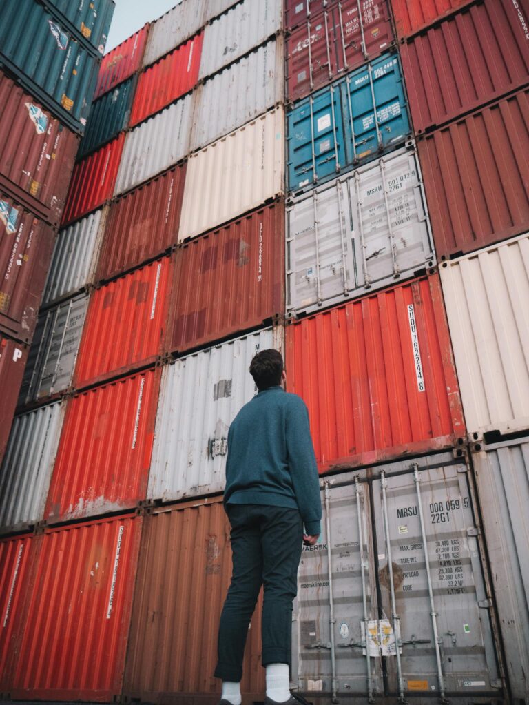 A man stands looking up at a stack of colorful cargo containers at a port, capturing the essence of global trade and commerce.